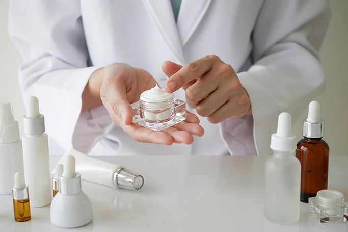 Una mujer con bata enseña una crema hidratante y otros productos de dermocosmética y cuidado de la piel